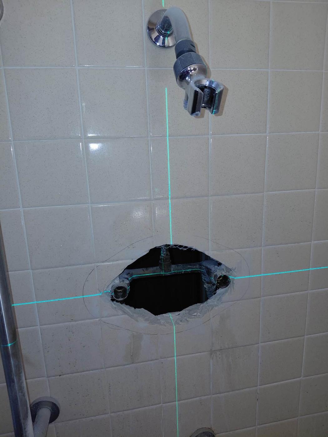 An access hole cut in shower tile with laser layout lines still marked on the wall